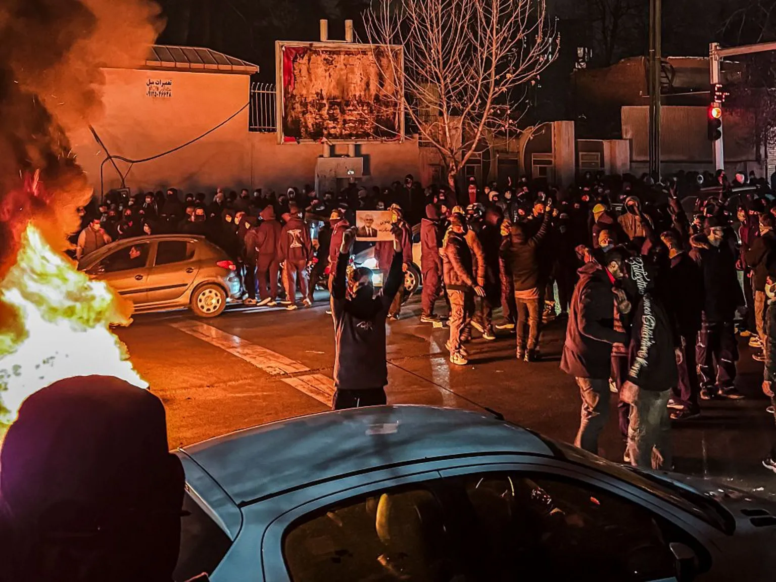Iranian protesters fill a street at night around a burning barricade, one holding a portrait aloft.