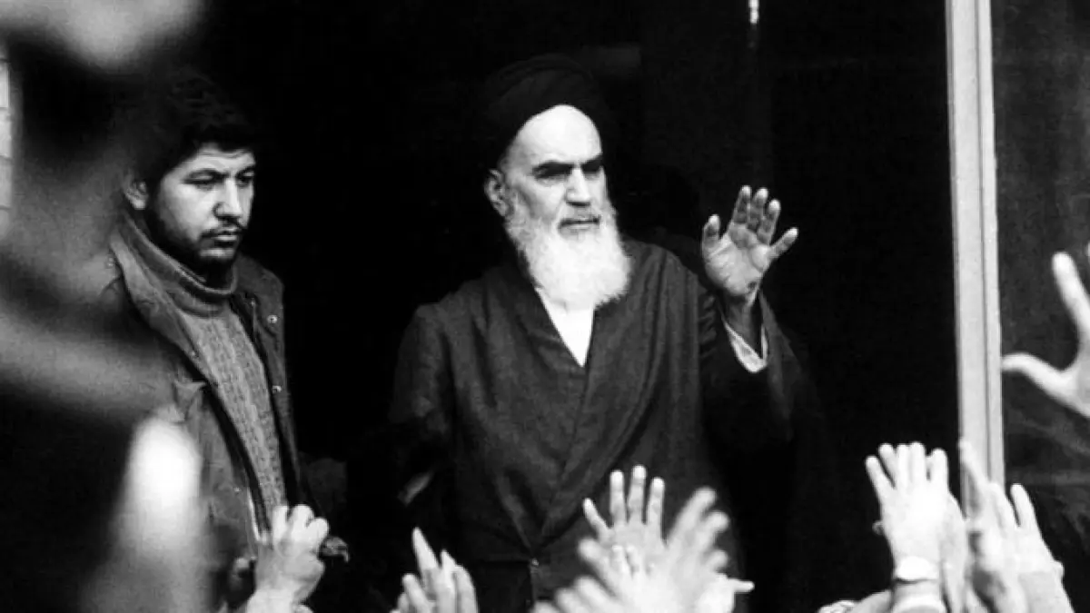 Ruhollah Khomeini raises his hand to a crowd reaching toward him, in a black-and-white photograph.
